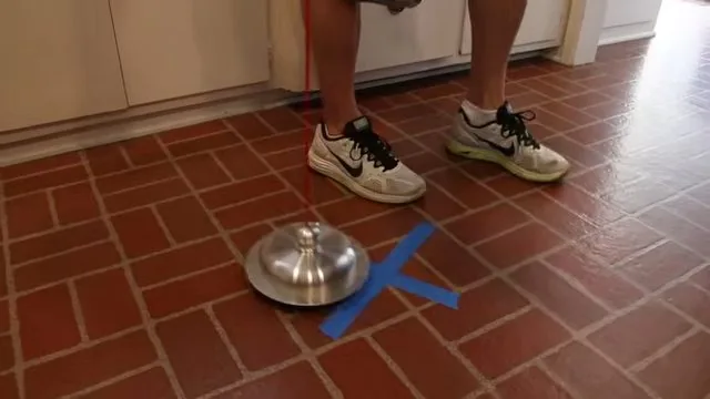 Metal listening disc resting on a tile floor beside blue tape in an X, with a person standing nearby