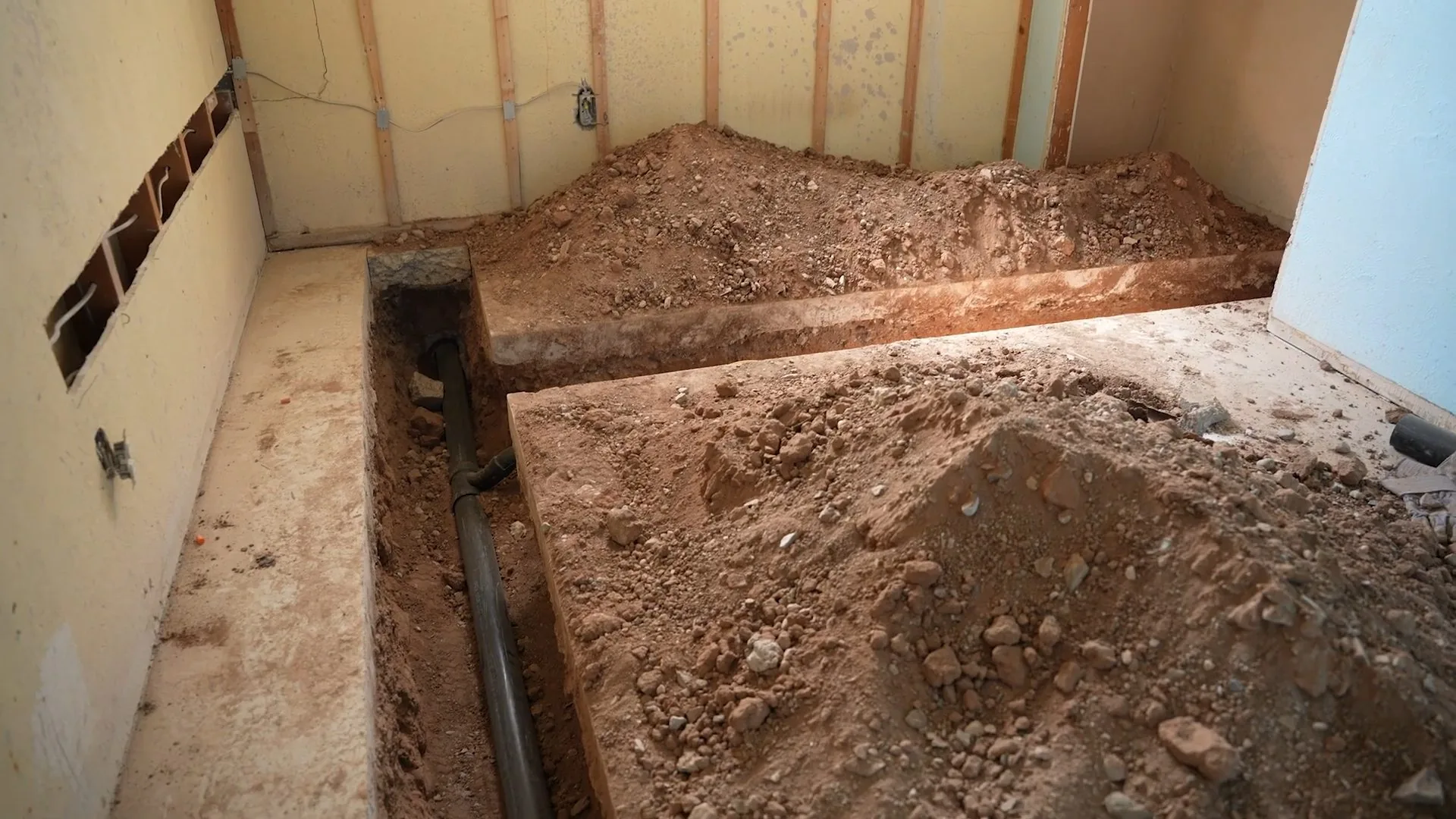 Basement floor opened along a framed wall, with a black drain pipe visible in the trench and piles of soil on either side