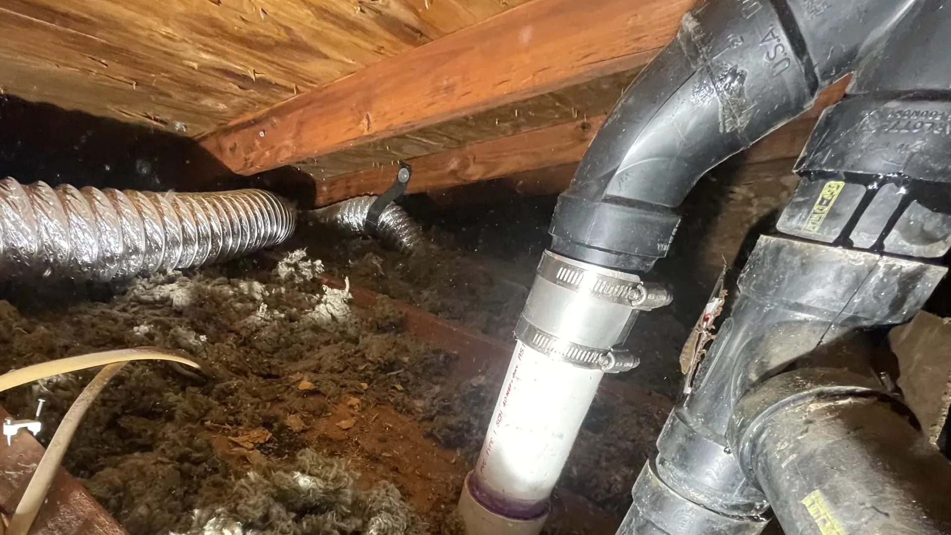 Black drain fittings and a white pipe joined with a steel clamp beneath wood joists in a crawlspace