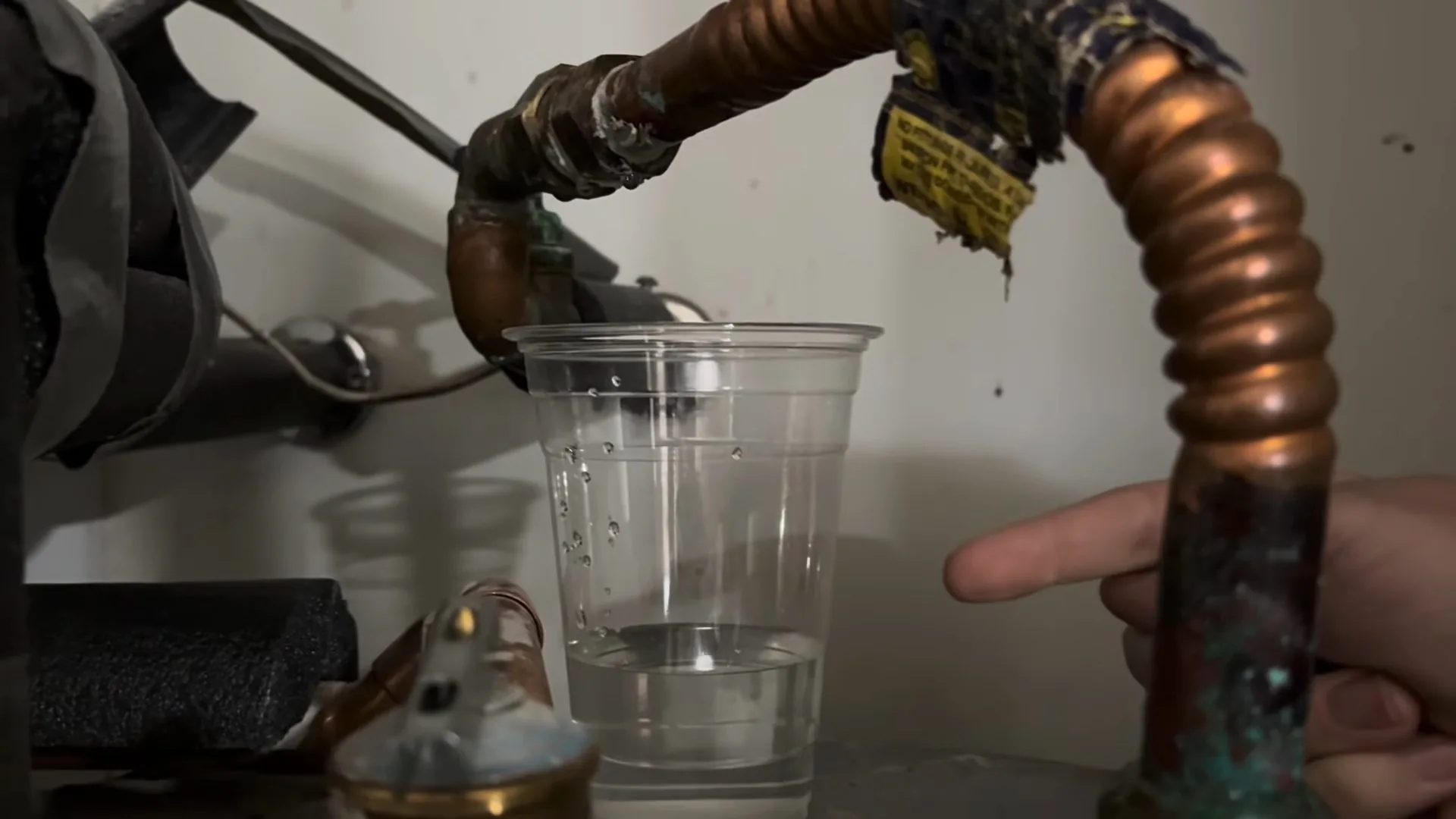 Corroded copper flex connector above a clear cup of water with a finger pointing at the drip