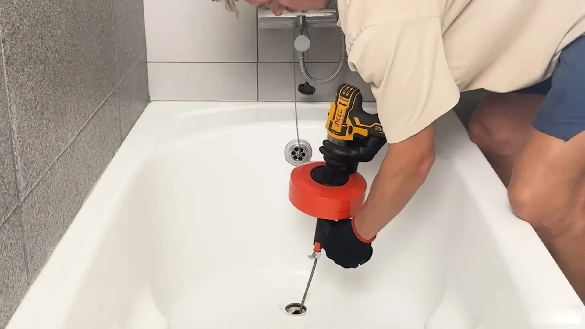 Person in a tan shirt feeding a small drain auger driven by a cordless drill into a white bathtub drain