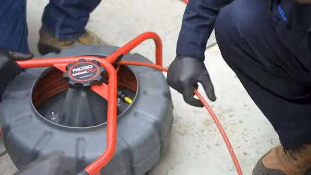 Two gloved technicians beside a red and gray drain cable machine with its cable running out across a concrete driveway
