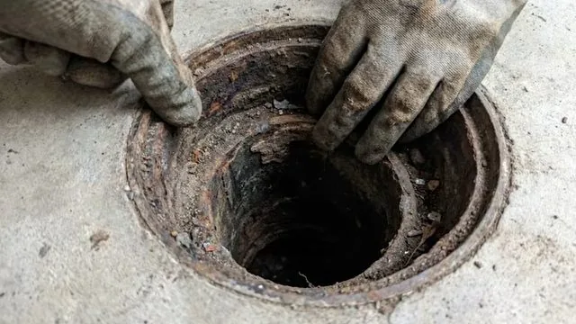 Two gloved hands at the rusted opening of a floor drain in a concrete floor