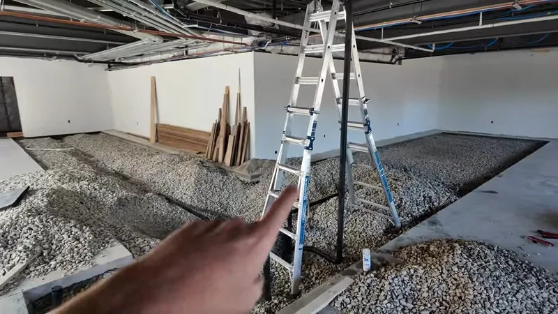 Gravel bed spread across an open floor area with a ladder, lumber and a hand pointing at the drain layout
