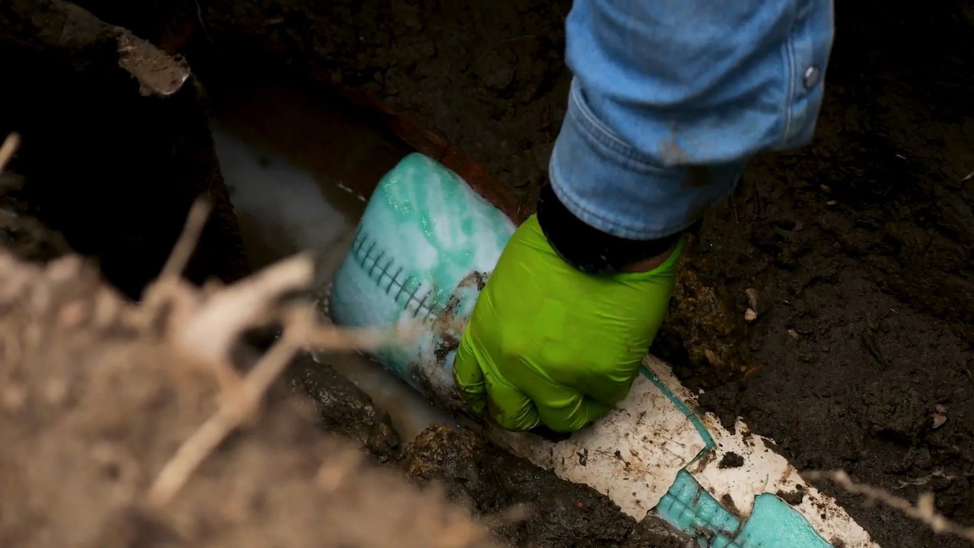 Green-gloved hand pressing a teal liner sleeve against a white pipe inside a soil trench with roots