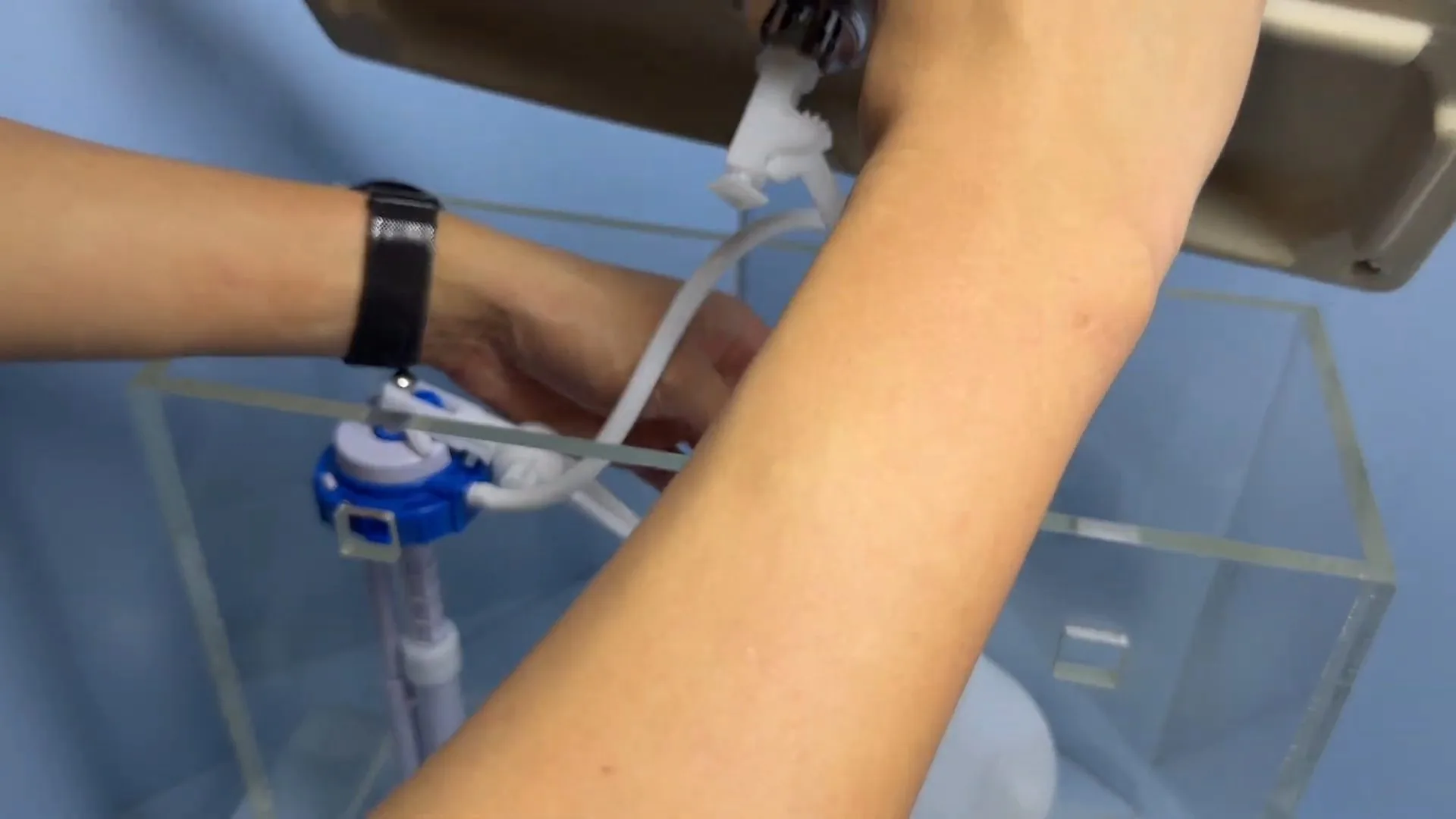 Forearms reaching into a clear acrylic toilet tank to adjust a blue fill valve