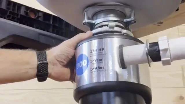 Hand gripping a stainless garbage disposal mounted under a sink with a white drain pipe attached