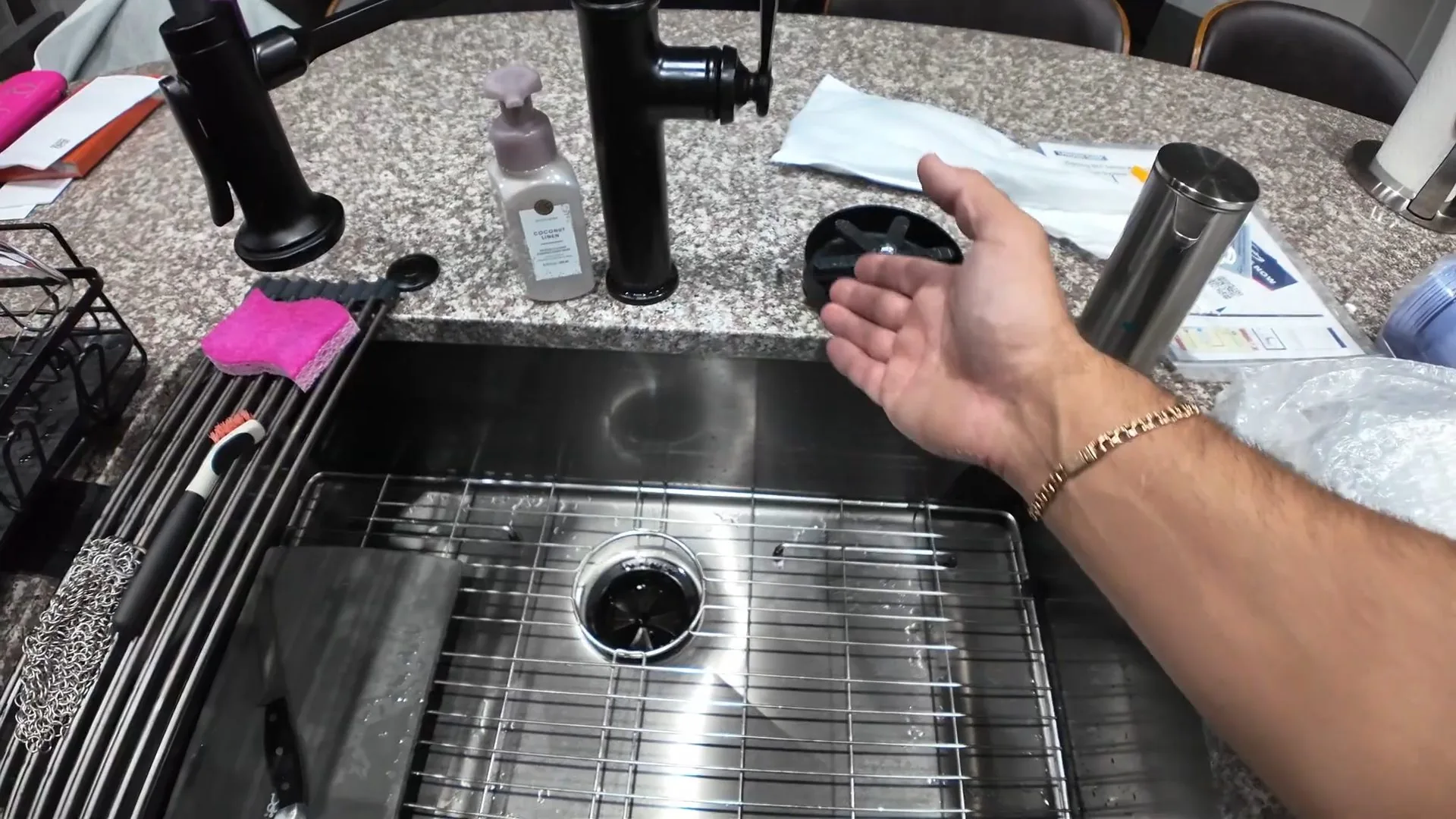 Hand held over a stainless steel sink with a drain opening, black faucets behind it and a granite counter