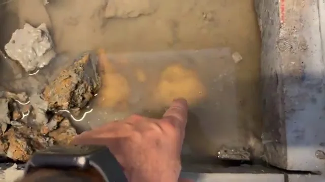 Hand pointing at a wet orange patch on an opened concrete slab beside broken rock