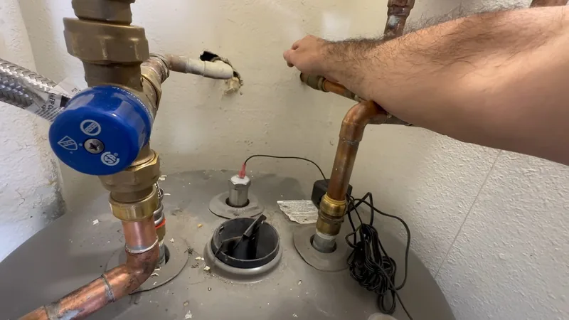 Arm reaching across the top of a water heater toward a blue-handled supply valve and copper pipes
