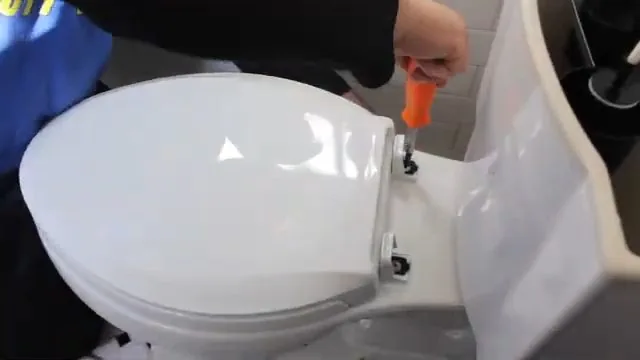 Hand using an orange screwdriver on the hinge of a white toilet seat