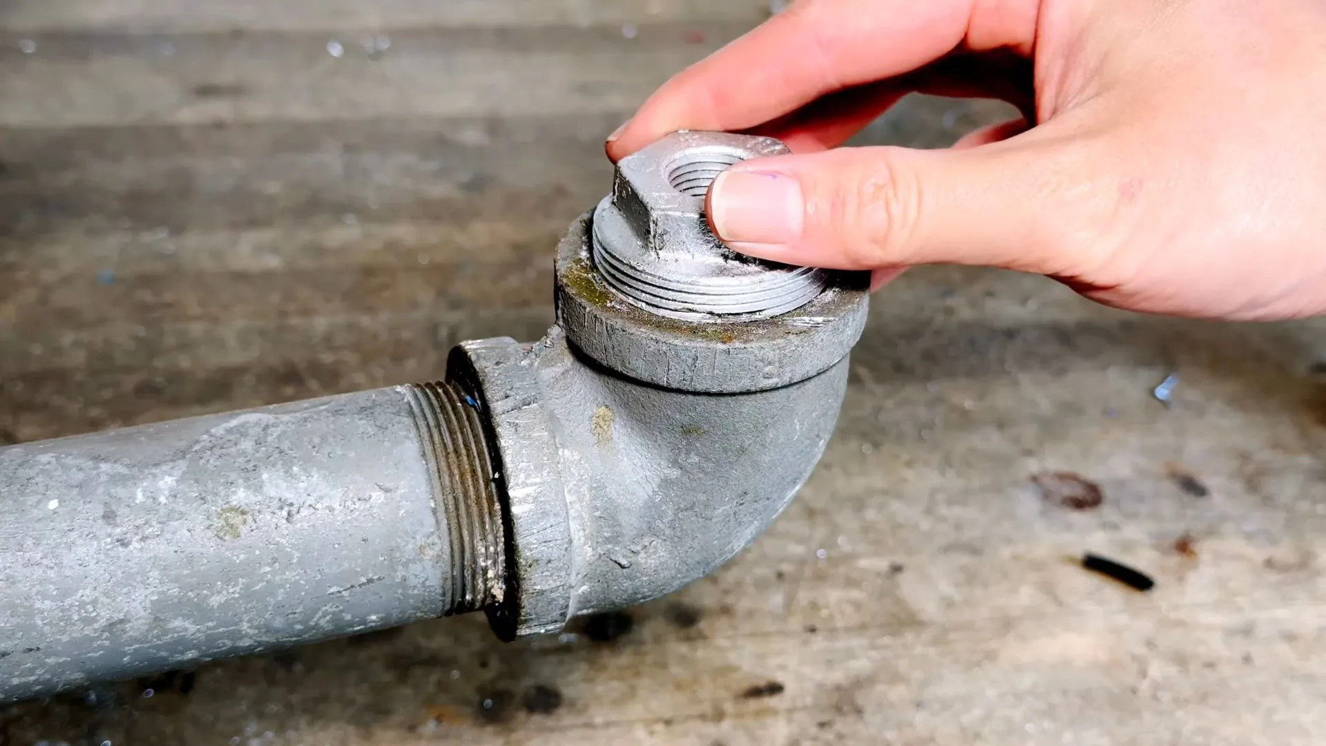Hand unscrewing the threaded cap of a galvanized cleanout fitting on a pipe lying on a concrete floor