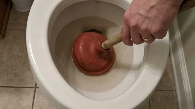 Hand pushing a red plunger down into a white toilet bowl on a tile floor