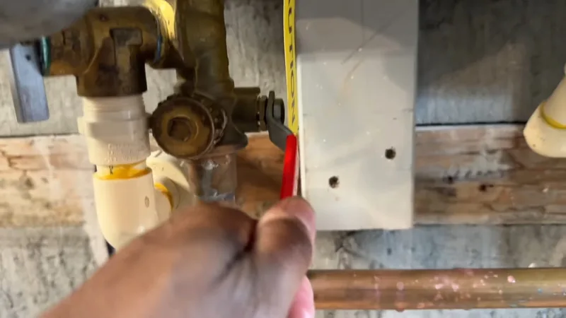 Hand holding a tape measure beside brass valves and copper pipes mounted on a basement wall