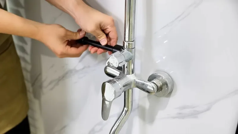 Hands using a small black tool on a chrome shower faucet mounted on a white marble wall