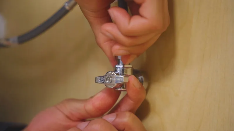 Hands working a chrome angle stop valve on a wooden cabinet wall