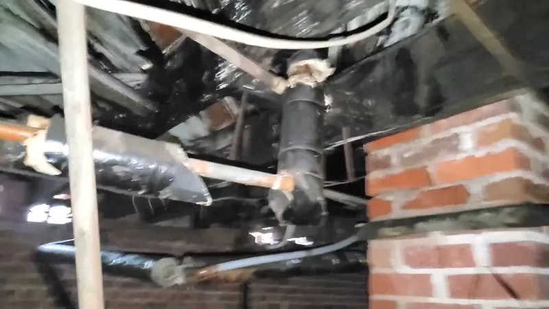 Foil-wrapped pipes and a black drain line running overhead in a brick-walled crawlspace