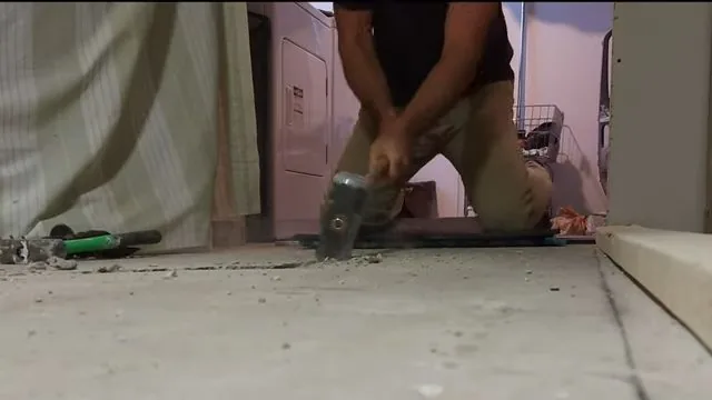 Kneeling man swinging a hammer to break a concrete floor beside a laundry area