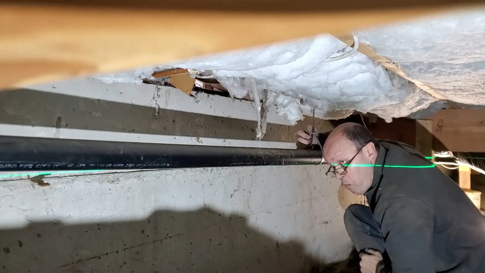 Man crouching beside a black pipe running under insulated floor joists with a green laser line across it