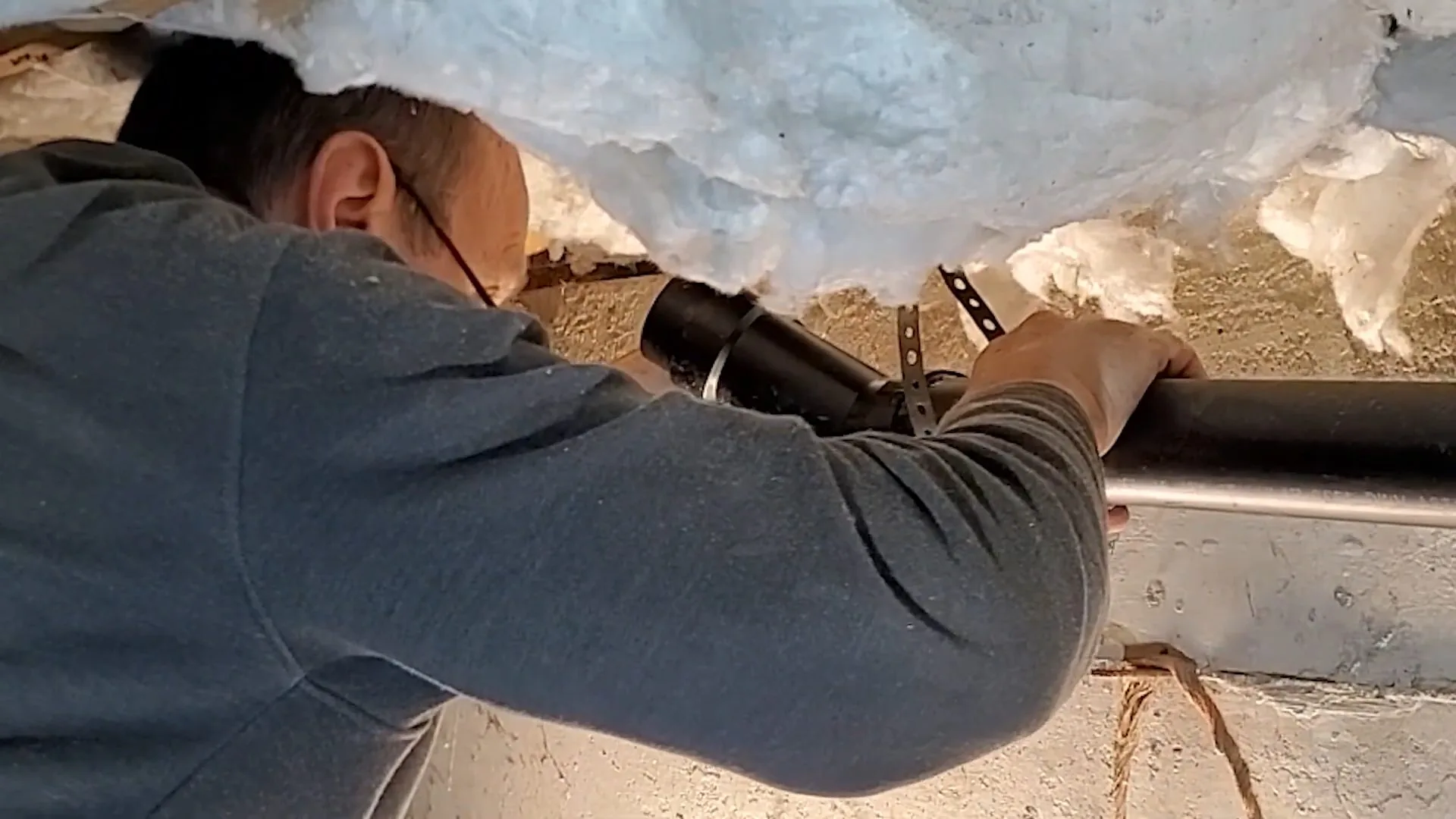 Man with glasses reaching overhead to fasten black pipe beneath torn fiberglass insulation