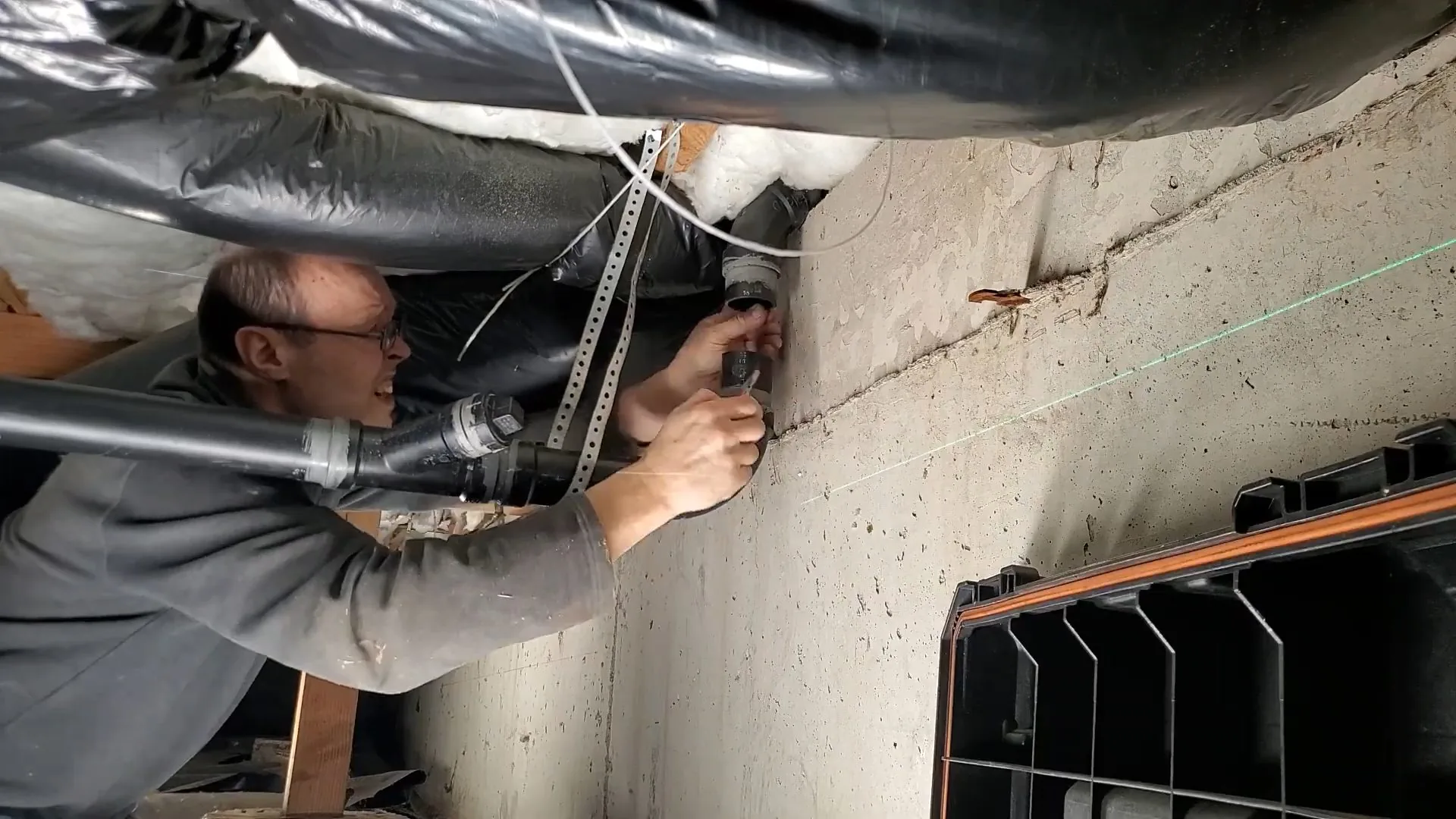Man in a gray sweatshirt reaching up to a black pipe fastened to a concrete basement wall