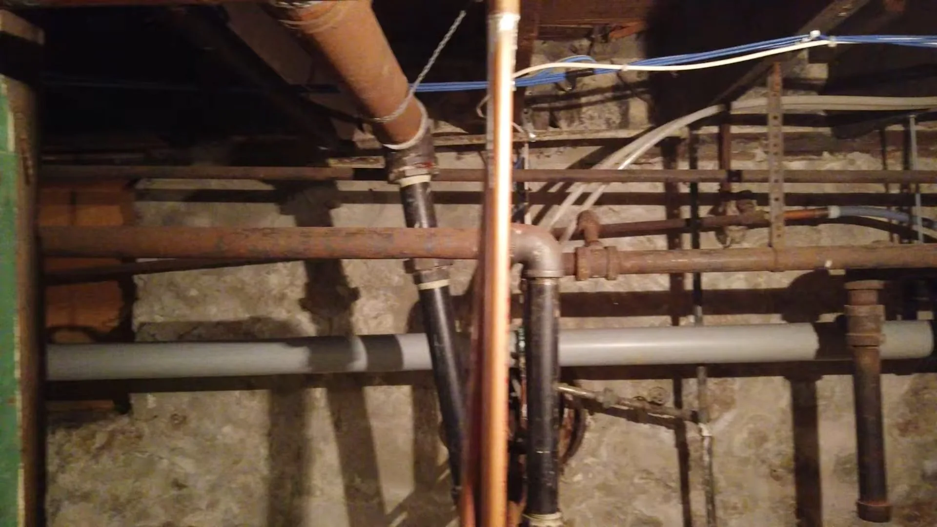 Mix of copper, iron and gray plastic pipes running below floor joists in an older basement