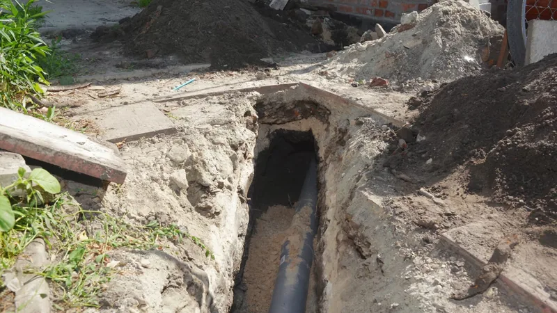 Open trench in a sandy yard with a black sewer pipe visible at the bottom