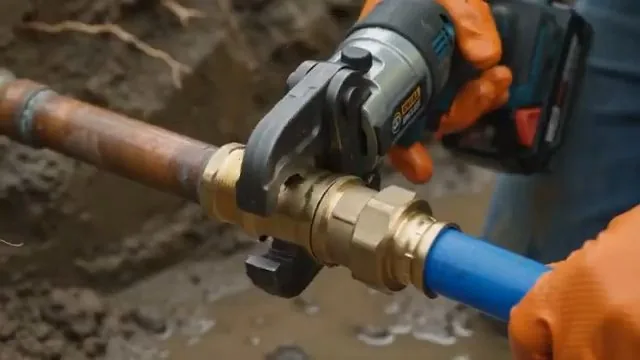 Orange-gloved hands using a battery crimp tool to join a brass valve to blue pipe in a dirt trench