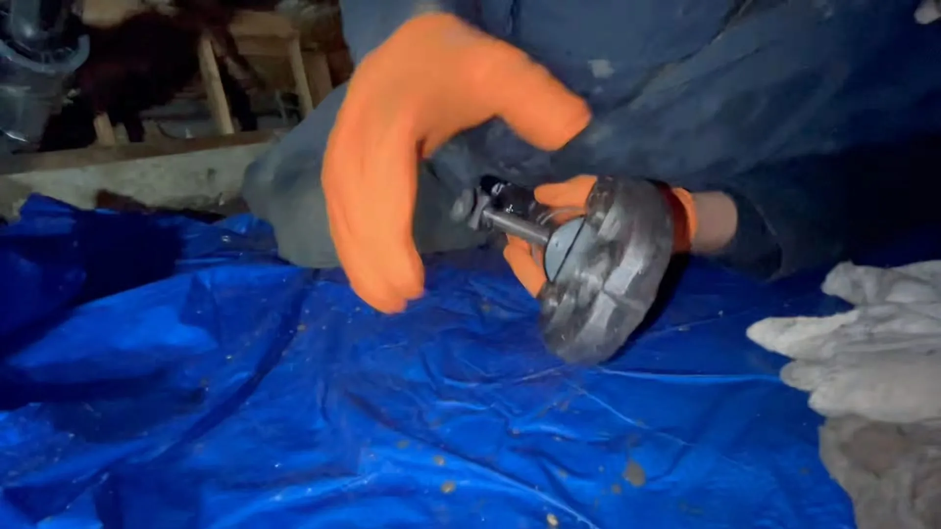 Orange-gloved hands holding a gray valve over a blue tarp in a crawlspace
