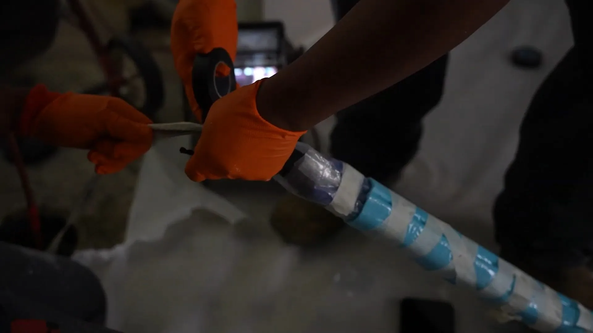 Orange-gloved hands wrapping blue tape around a drain camera cable while a monitor shows in the background