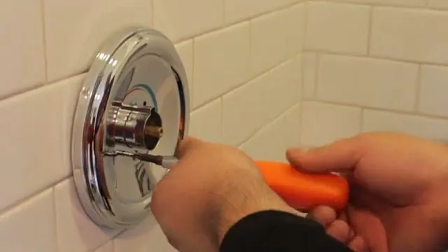 Hands holding an orange screwdriver at a chrome shower trim plate on a white tile wall
