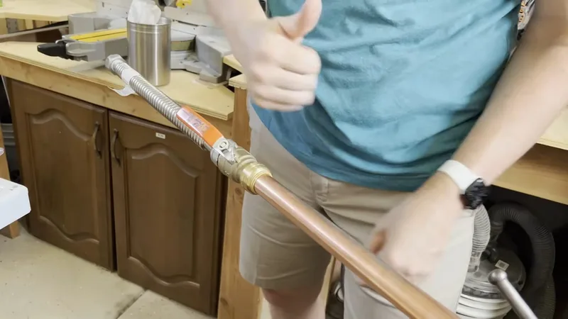 Person in a teal shirt holding a stainless flexible gas connector attached to a copper line, giving a thumbs up