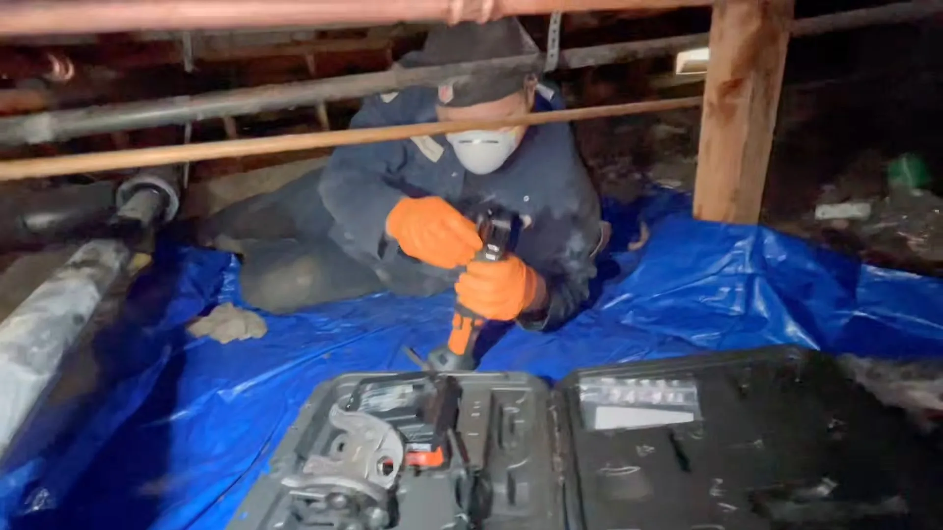 Plumber in a dust mask and orange gloves working on a pipe in a crawlspace, kneeling on a blue tarp