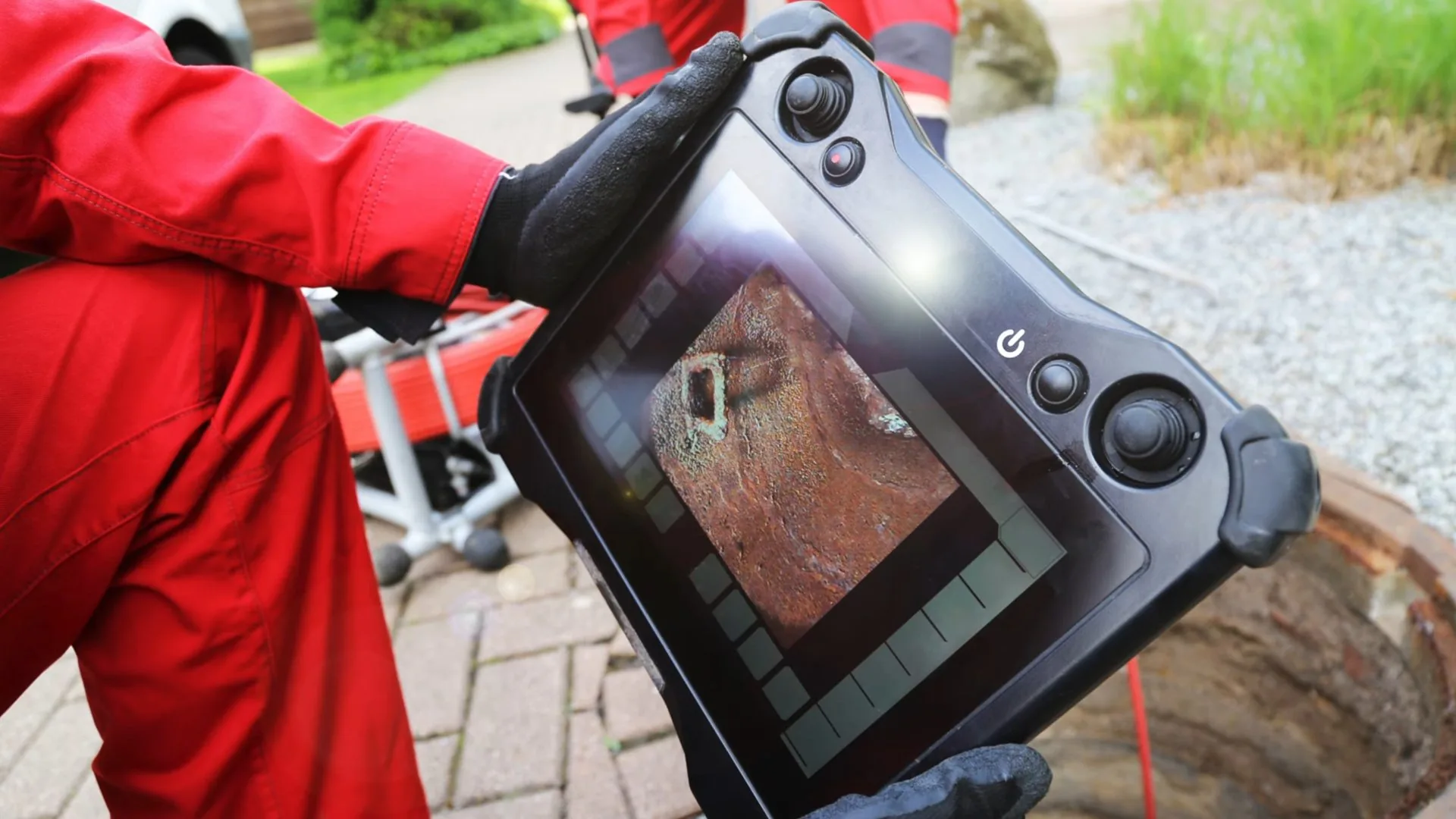 Technician in a red suit holding a camera control tablet beside a paved yard cleanout