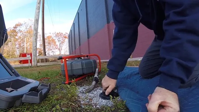 Technician in a navy jacket kneeling beside a lawn cleanout with a red camera reel and monitor case on the grass