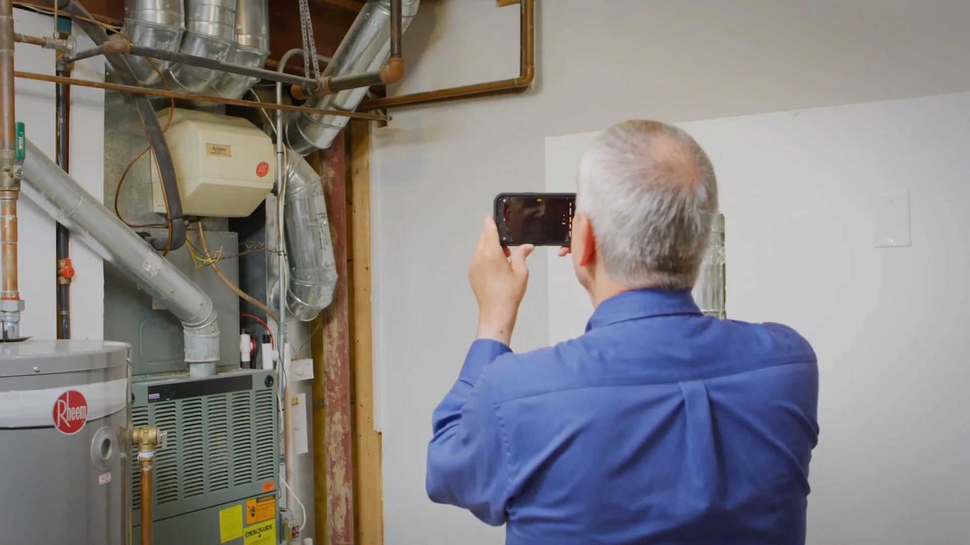 Technician in a blue shirt taking a phone photo of a basement wall beside a gas water heater and furnace