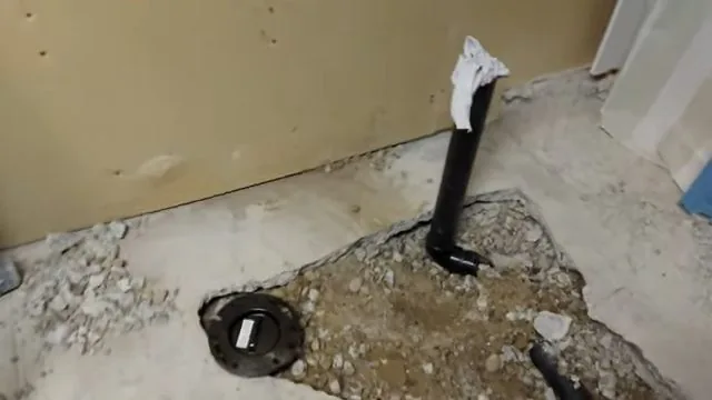 Black toilet flange set in a broken concrete floor next to a black drain pipe wrapped in white tape