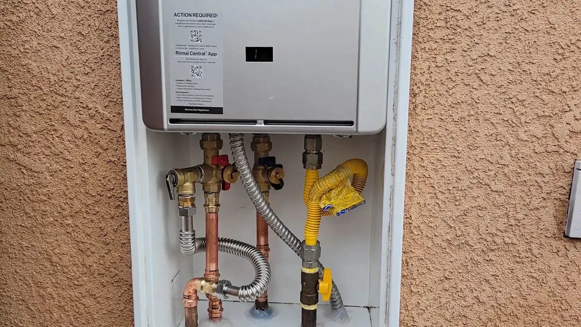 Wall-mounted tankless water heater in a recessed stucco alcove with copper water lines and a yellow gas line below it