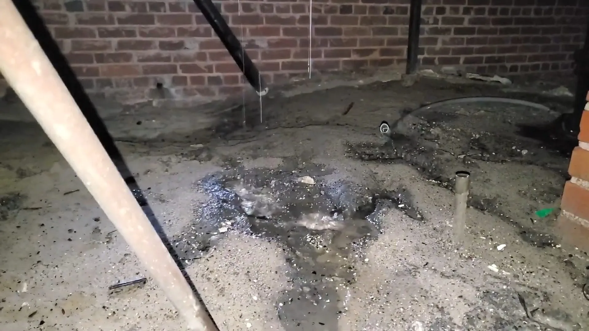 Wet concrete floor in a brick-walled basement with water dripping from a pipe above
