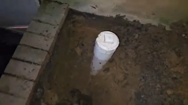 White capped cleanout pipe standing up from a dirt floor beside a brick wall