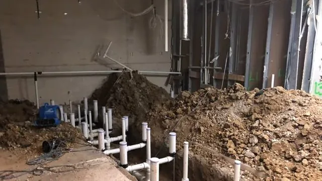 Row of white PVC drain stubs standing in an open trench in a basement floor beside piles of soil